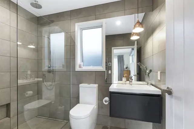 Grey tiled bathroom with glass shower, toilet and dark vanity