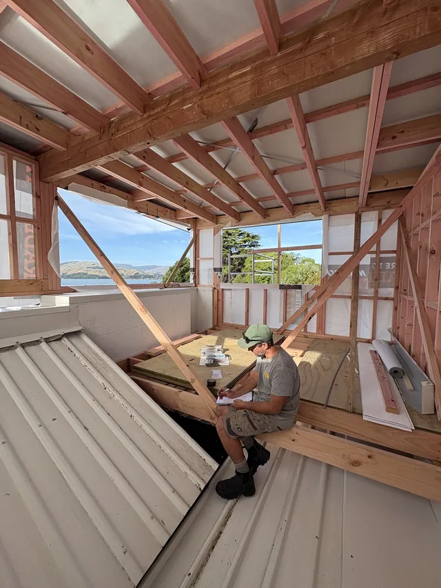 Builder sitting on a floor joist inside the framed new level, with hills and water visible through the openings