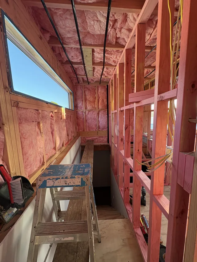Framed walls and ceiling with pink insulation and wiring in place, and a ladder at the top of the stairwell
