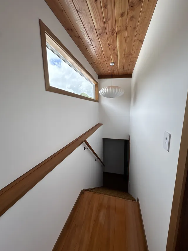 Looking down the stairwell from the top landing, with a timber floor, timber handrail, high window, pendant light and timber-lined ceiling