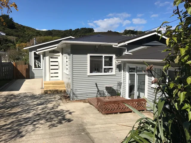 The house with grey weatherboards, white joinery and a dark roof, with timber front steps and a small deck with outdoor chairs, seen from the driveway
